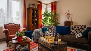 Ein gemütliches Wohnzimmer mit einem bequemen Ohrensessel, einer Couch mit gemusterten Kissen, einem Holzschrank und einer großen Pflanze.