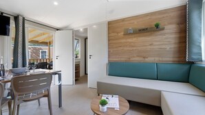 A modern living room with a sofa, a dining table with chairs, a small table with a plant, and a wall-mounted shelf with a sign. - “Evasion 55” Mobile Home with Shared Pool and Terrace (Saint-Gervais)