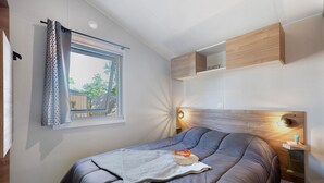 A modern bedroom with a large bed, a window with curtains, a bedside table, and a built-in shelf. - “Evasion 55” Mobile Home with Shared Pool and Terrace (Saint-Gervais)