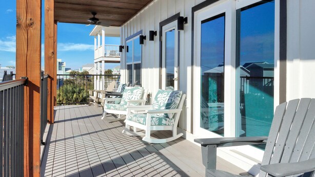 A covered deck with white rocking chairs and a ceiling fan, overlooking a pool and a building with large windows. - The Lazy Turtle (Port St. Joe)