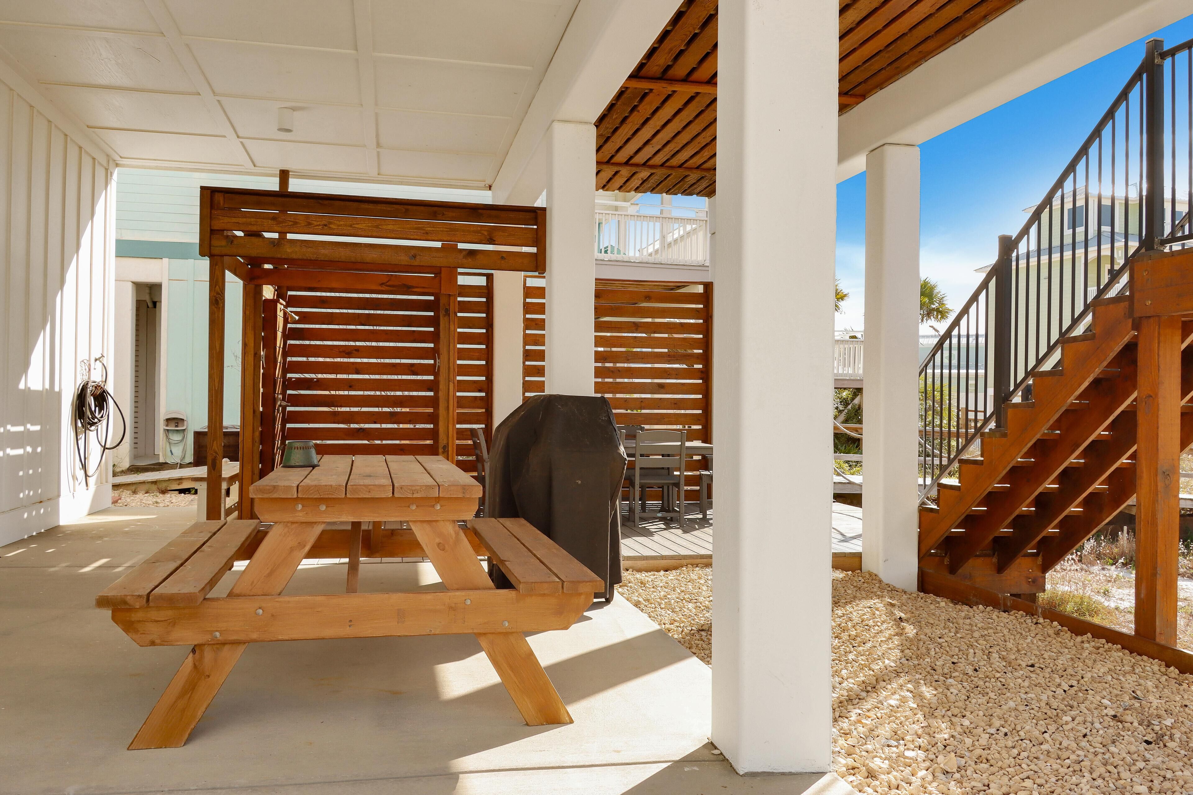 Un espacio exterior techado con una mesa de picnic de madera, una parrilla para barbacoa y una escalera que lleva a un nivel superior.
