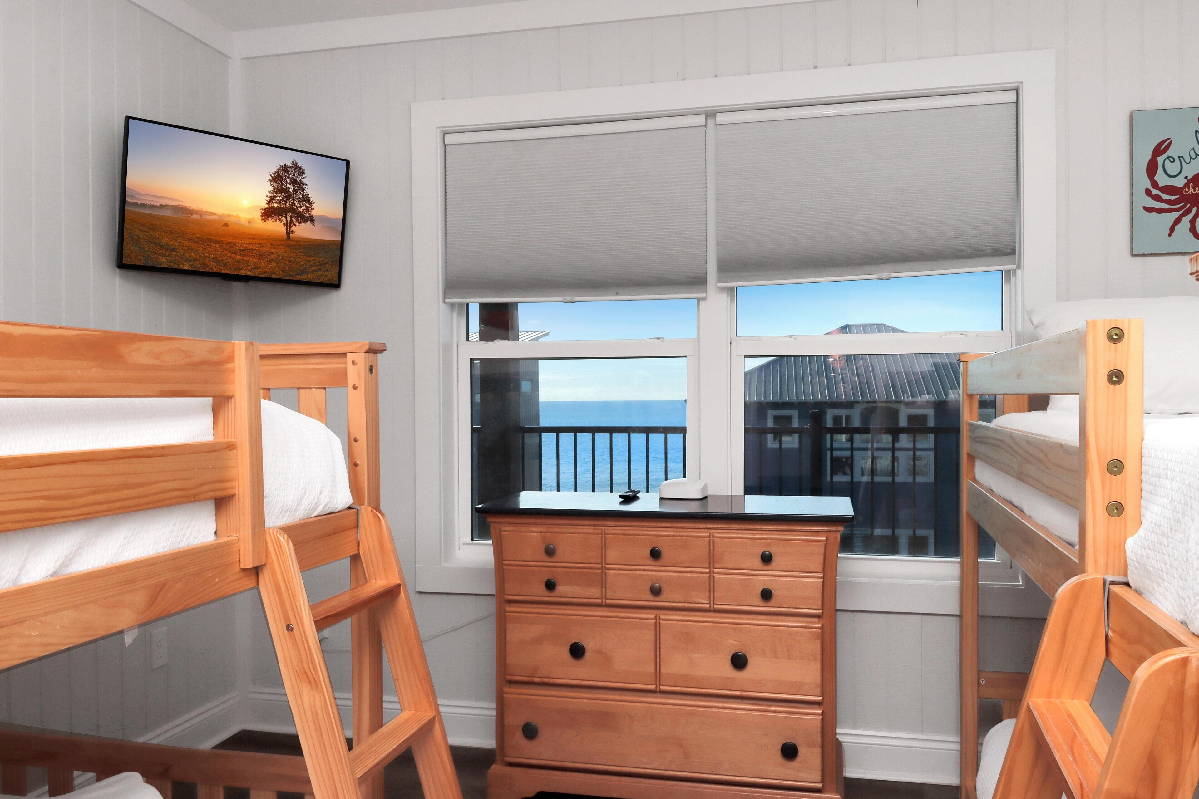 Ett sovrum med två våningssängar, en byrå i trä, en tv och ett fönster med utsikt över havet.