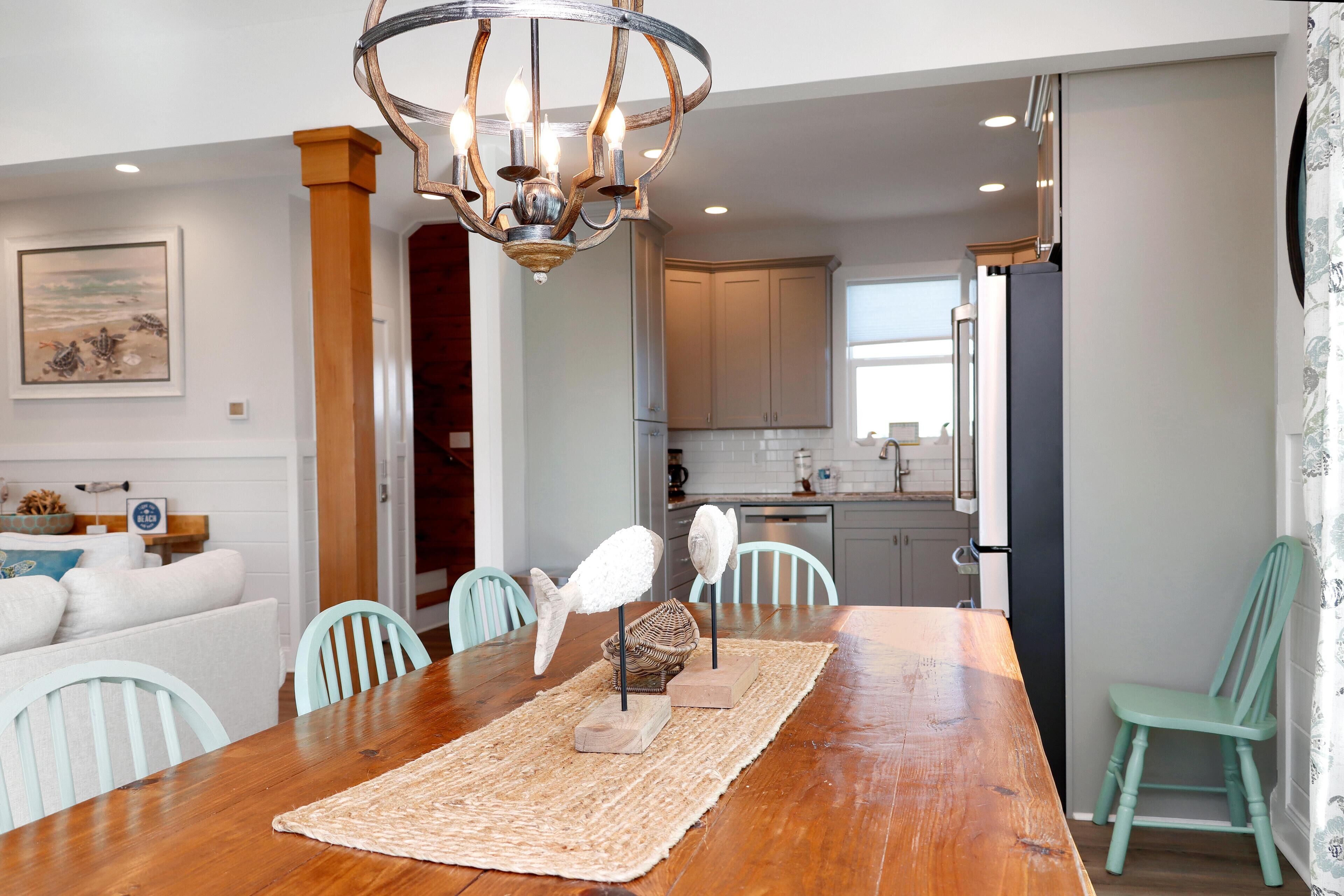 A dining area with a wooden table, chairs, a chandelier, and a kitchen in the background.