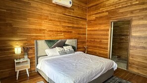 Une chambre avec un mur lambrissé en bois, un lit doté d’une tête de lit rembourrée de couleur grise, une table de chevet avec une lampe et une porte coulissante.