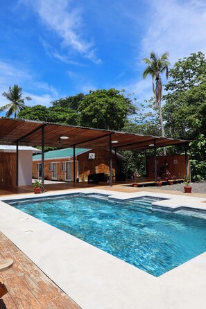 Une piscine avec une terrasse en bois, un patio couvert et un bâtiment à l’extérieur en briques.