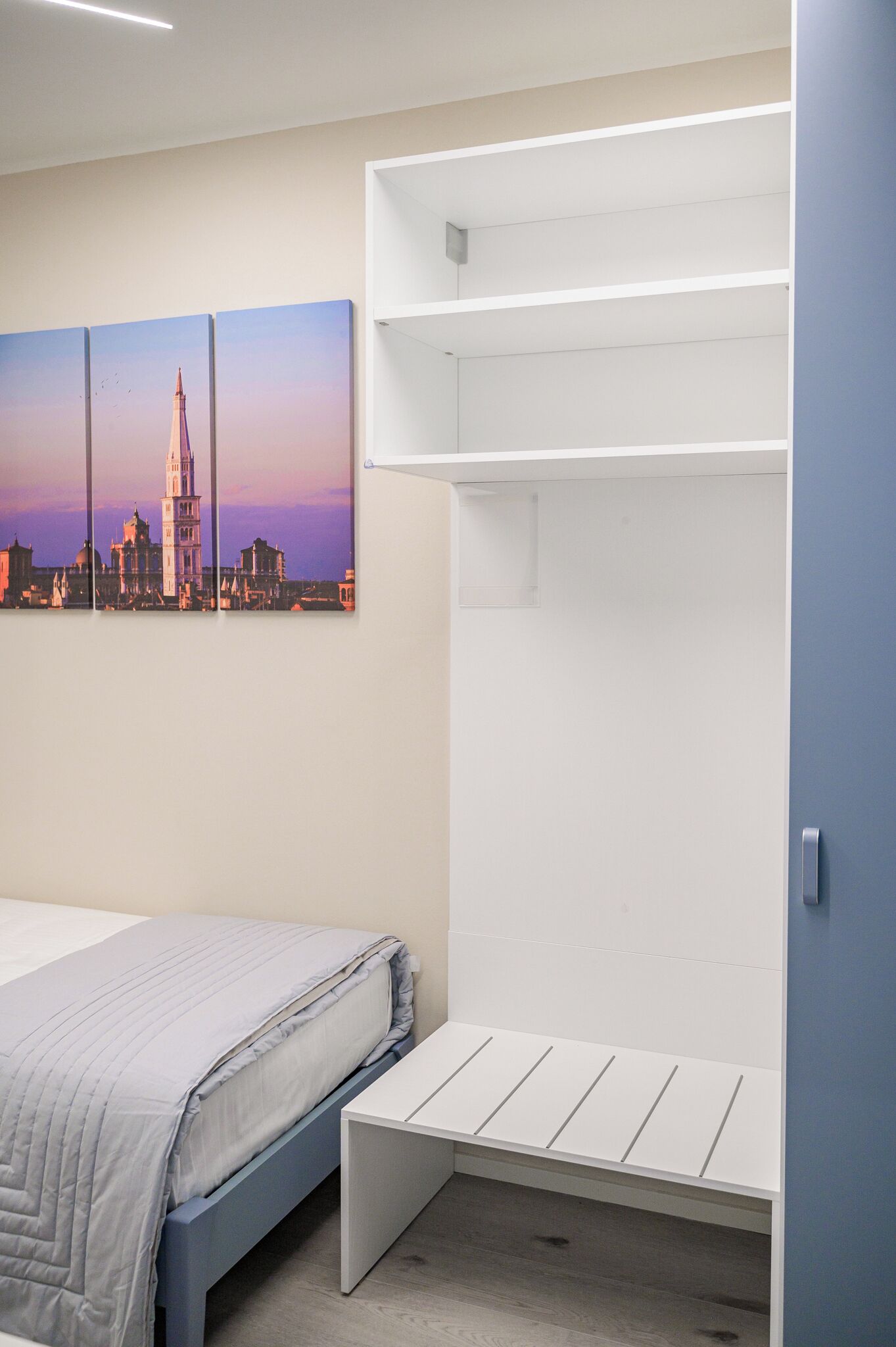 A bedroom with a bed, a white bench, a wall-mounted shelf, and a cityscape painting.