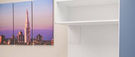 A bedroom with a bed, a white bench, a wall-mounted shelf, and a cityscape painting.