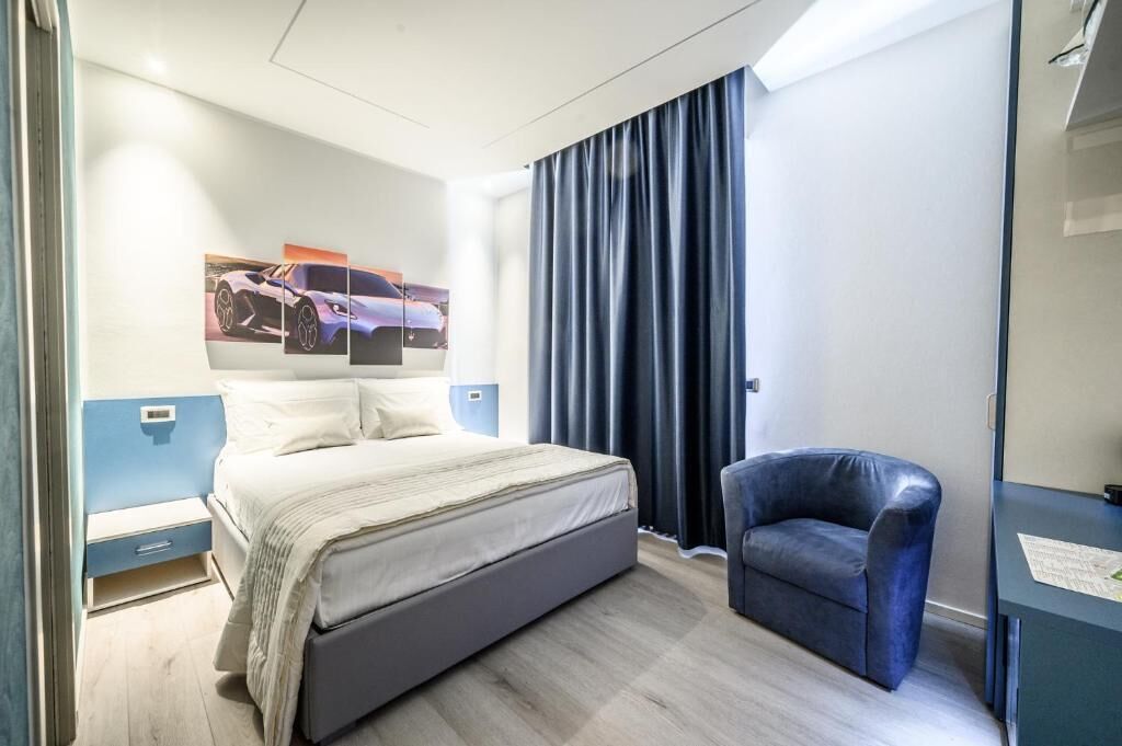 Ett modernt hotellrum med en stor säng, en blå fåtölj, ett skrivbord med en lampa och ett konstverk föreställande en bil som hänger på väggen.