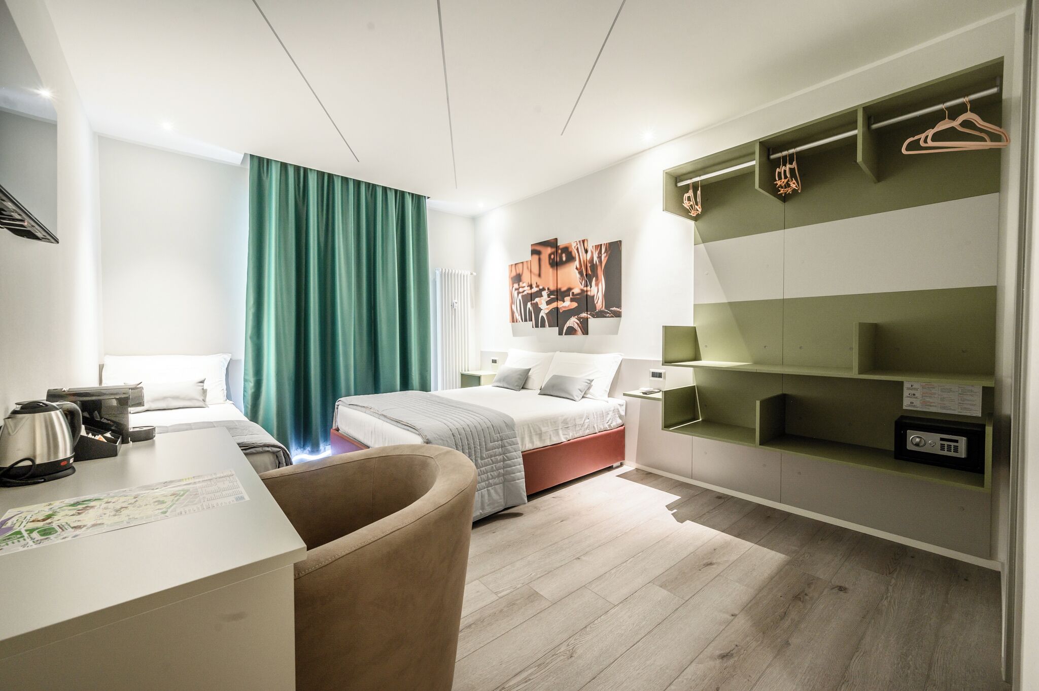 A modern hotel room with a bed, a desk, a chair, a green curtain, a wall-mounted TV, and a shelf with hangers.