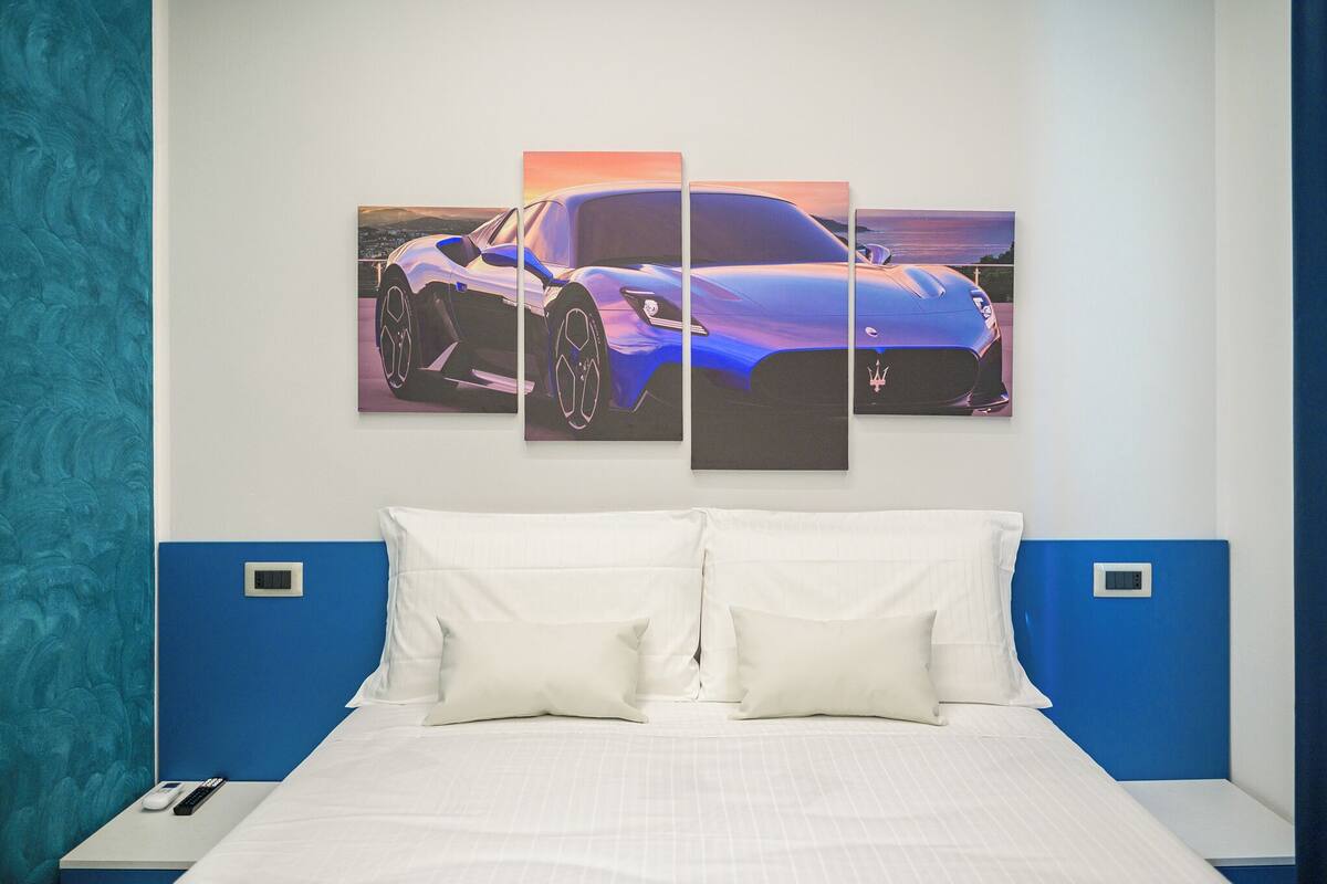 A hotel room with a large bed, white bedding, a blue headboard, and a wall-mounted artwork of a blue sports car.