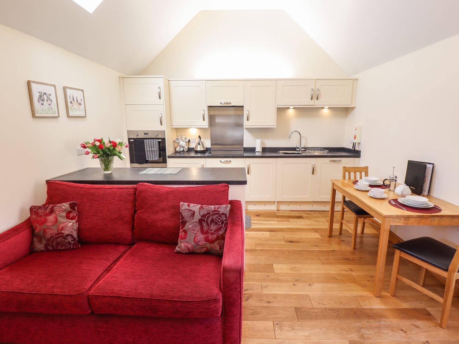 A compact living space with a kitchenette, a red sofa with floral cushions, a dining table with chairs, and a small TV.