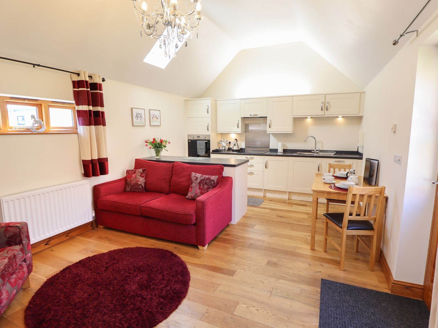 A compact living space with a red sofa, a dining table with chairs, a kitchen area, and a chandelier.