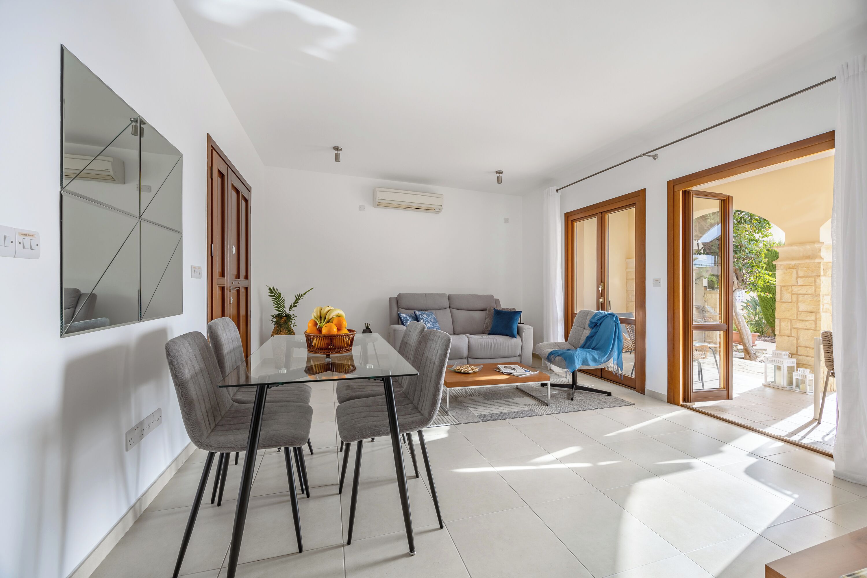A modern living room with a glass dining table, grey sofas, and a large mirror.