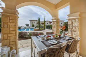 Uma área de refeições ao ar livre com pilares de pedra, uma mesa posta para dois e vista para uma piscina e edifícios ao fundo.