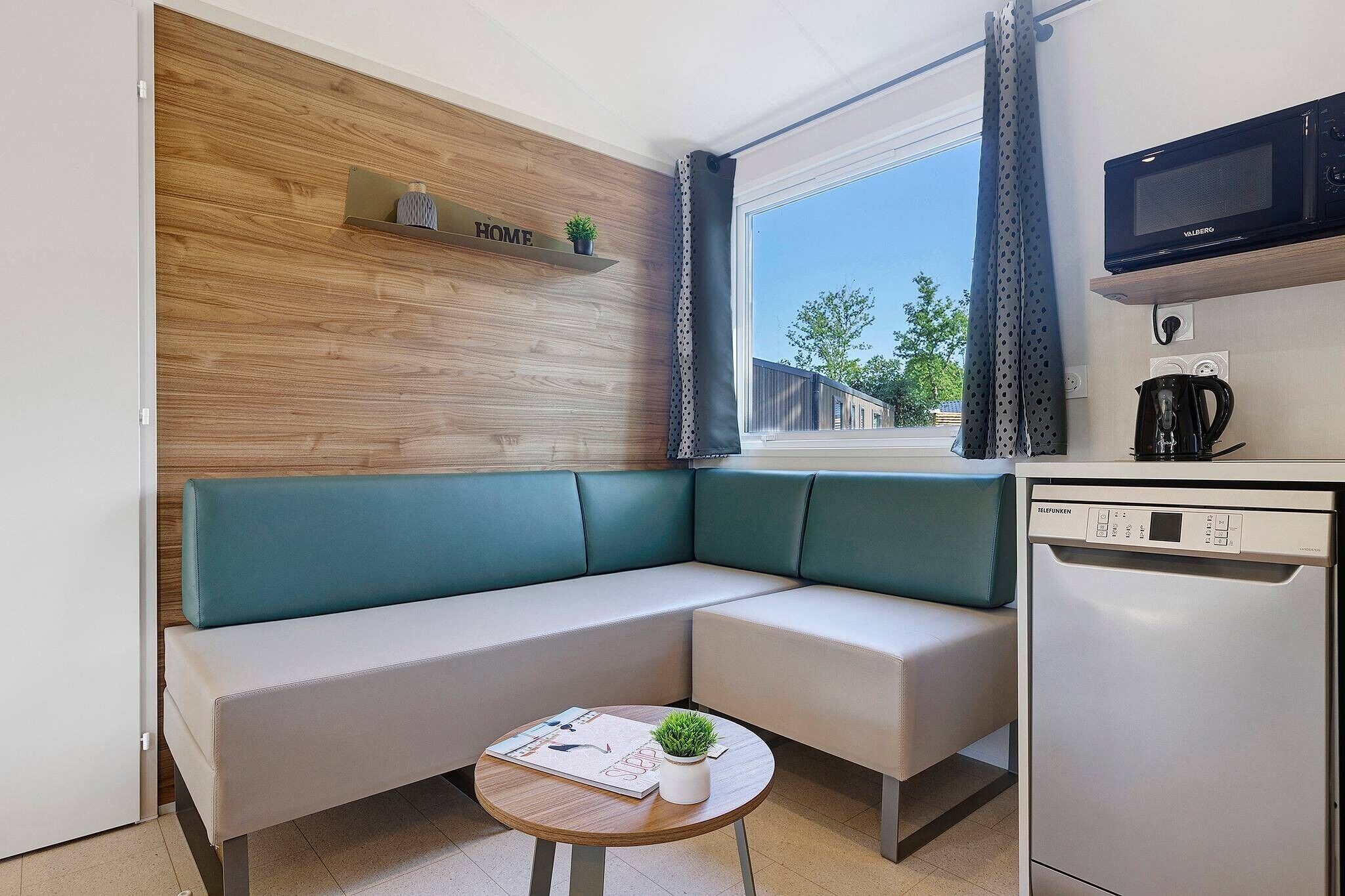 A modern kitchenette with a microwave, coffee maker, and dishwasher. A green sofa and a small round table with a magazine and a plant are also visible.