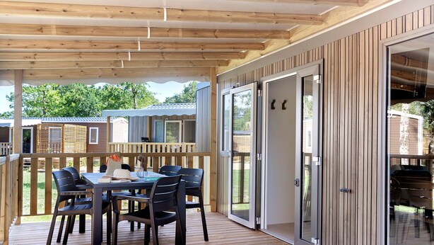 A covered outdoor area with a wooden deck, a table with chairs, and a sliding glass door leading to an interior space. - “Evasion 52” Mobile Home with Shared Pool and Terrace (Saint-Gervais)