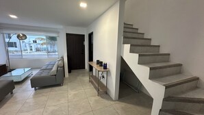 A modern living room with a grey sofa, a glass coffee table, and a wooden side table with two bottles. A staircase with grey steps leads to an upper floor. - Blue Villas by Host Ecuador (Santa Elena)