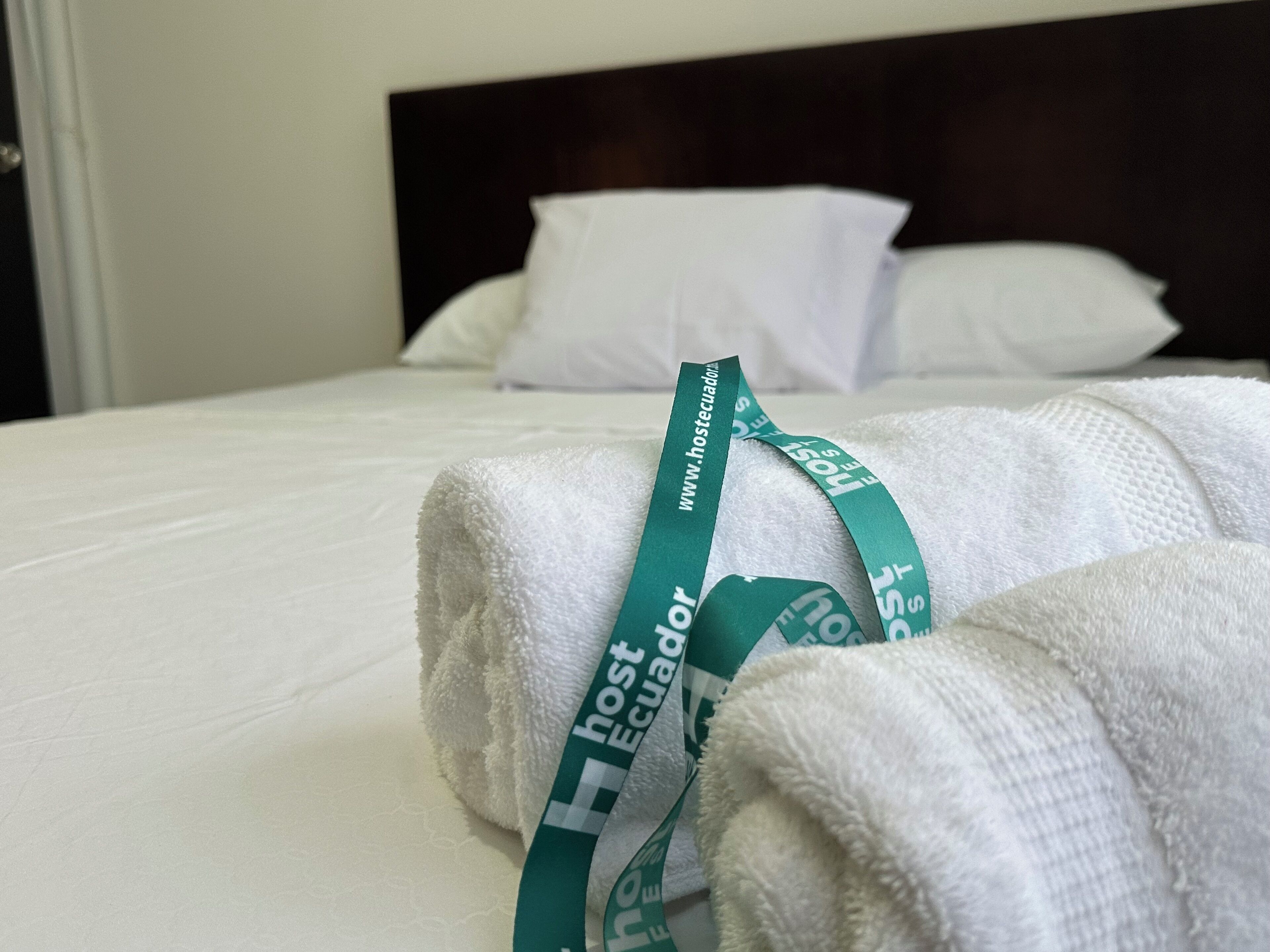 A neatly made bed with white linens and a green hotel towel with the text "hostecuador.com".