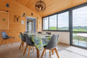 A dining area with a table set for four, wooden walls, and a large window with a view. - La Maison Bleue du Plot – Peaceful Stone House with Pool and Garden (Ailhon)