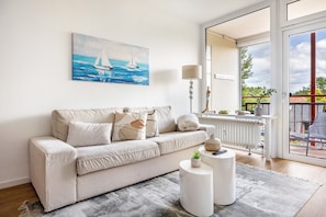 A living room with a beige sofa, a painting of sailboats, a lamp, and a view through a glass door. - Luxurious Small Coastal Nest Apartment Directly on Lake Steinhude (Wunstorf)
