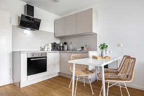 A compact kitchen with a built-in oven, a dining table with two wicker chairs, and a small potted plant. - Luxurious Small Coastal Nest Apartment Directly on Lake Steinhude (Wunstorf)