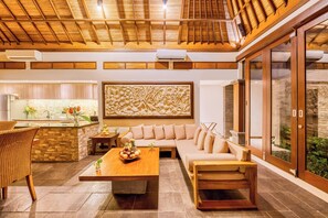 A modern living room with a wooden ceiling, a large wall art piece, a sofa, a coffee table, and a kitchen area with a stone backsplash. - Villa Jalamati - 3 BDR Luxury Escape, Prime Location (Kerobokan Kelod, Kec. Kuta Utara, Kabupaten Badung)