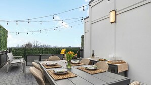 Une terrasse sur le toit avec une table à manger pour quatre personnes, entourée de chaises en osier, d’une petite table ornée d’un vase de fleurs et de guirlandes lumineuses.