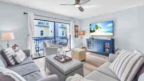 Un soggiorno moderno con una grande televisione, posti a sedere comodi, un balcone vista edifici e una ventola a soffitto.