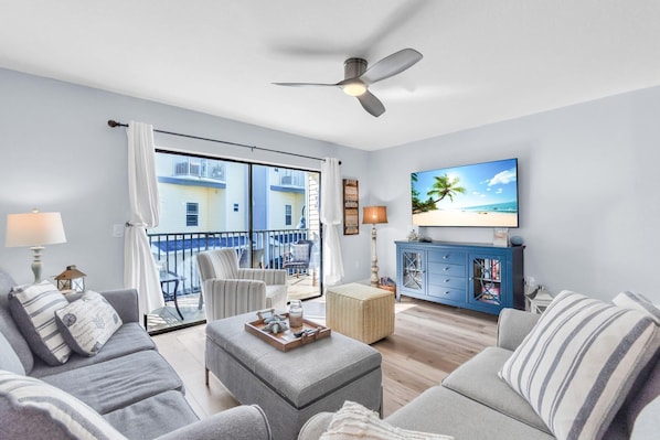 A modern living room with a large TV, comfortable seating, a balcony with a view of buildings, and a ceiling fan. - Bayview Villas - Bayview Shores (Indian Shores)