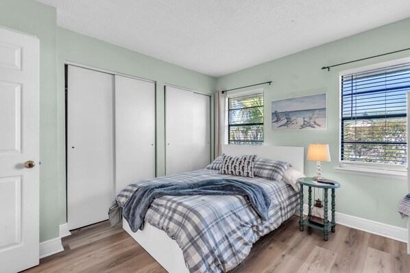 A bedroom with a bed, a nightstand, a window with blinds, and a door. - Bayview Villas - Bayview Shores (Indian Shores)