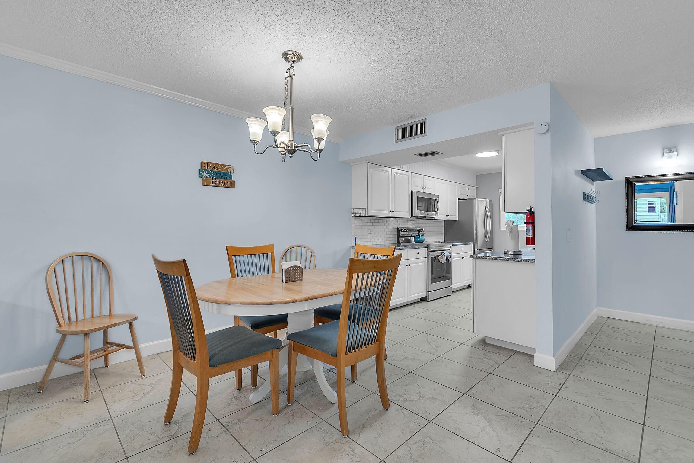 A dining area with a round table and four chairs, a chandelier, and a kitchen in the background.