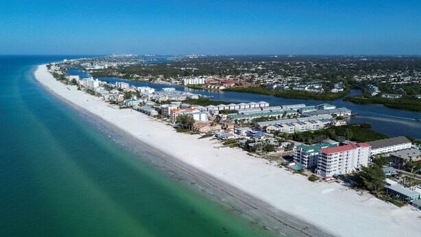 An aerial view of a coastal area with a long sandy beach, a marina with boats, and a mix of residential and commercial buildings. - Bayview Villas 120 (Indian Shores)