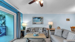 A living room with a blue wall, a ceiling fan, a painting of a beach, a coffee table with a bowl, and a grey sofa. - Bayview Villas 120 (Indian Shores)