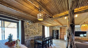 Un interior rústico con paredes de piedra, vigas de madera en el techo, un comedor con sillas, un televisor y un ventanal grande.