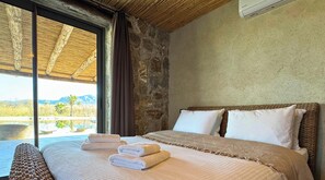A bedroom with a large bed, two pillows, a window with a view of mountains, and a stone wall. - Villa Zer – Unique Lagoon-Style Pool by Rental Finders Dalyan (Dalyan)
