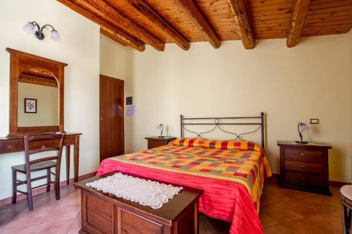 A bedroom with a wooden ceiling, a bed with a colorful quilt, a desk with a chair, and a nightstand with a lamp.