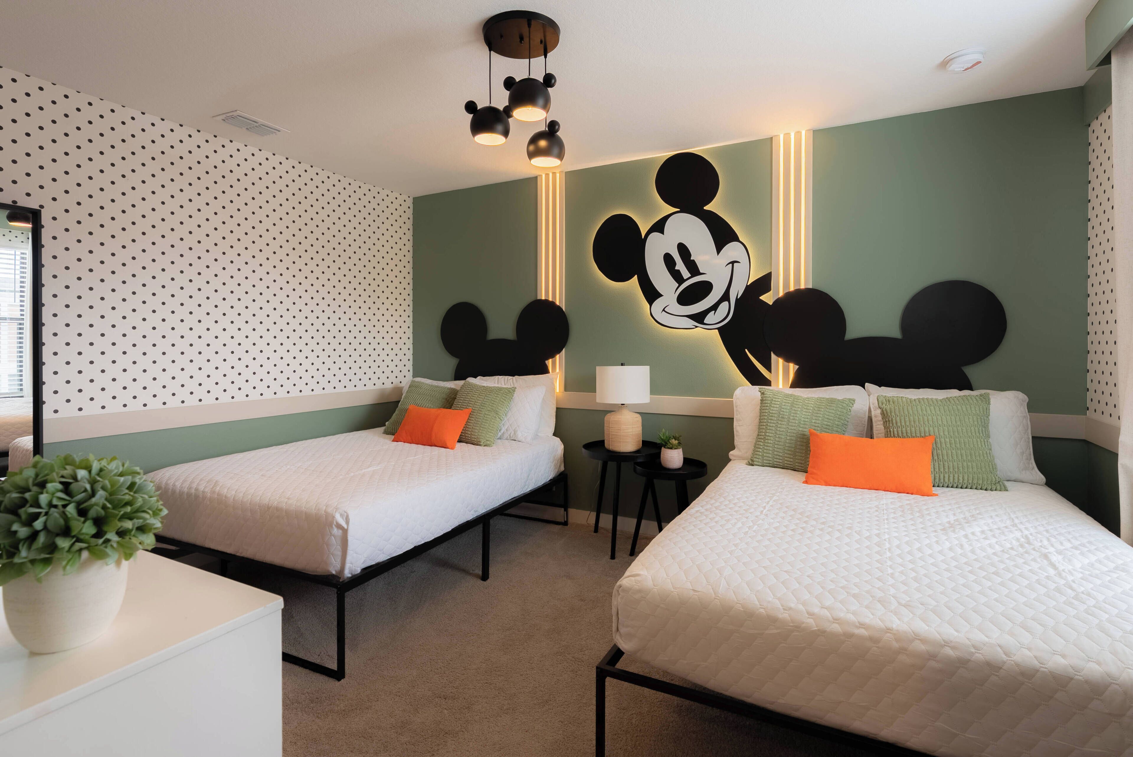 A hotel room with two beds, a Mickey Mouse mural, a small table with a lamp, and a potted plant.