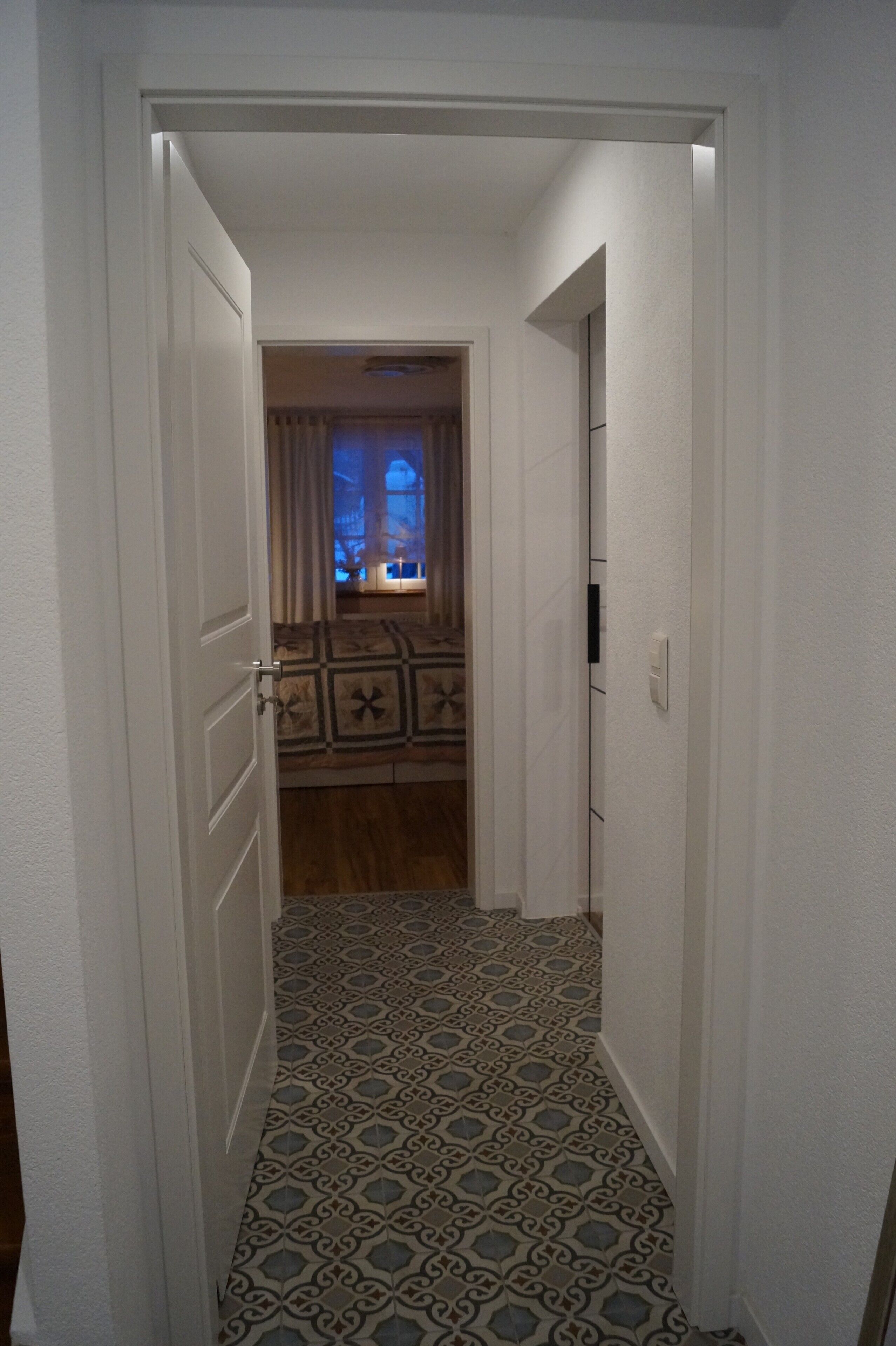 A narrow hallway with patterned flooring, a door on the left, and a room with a bed and curtains in the background.