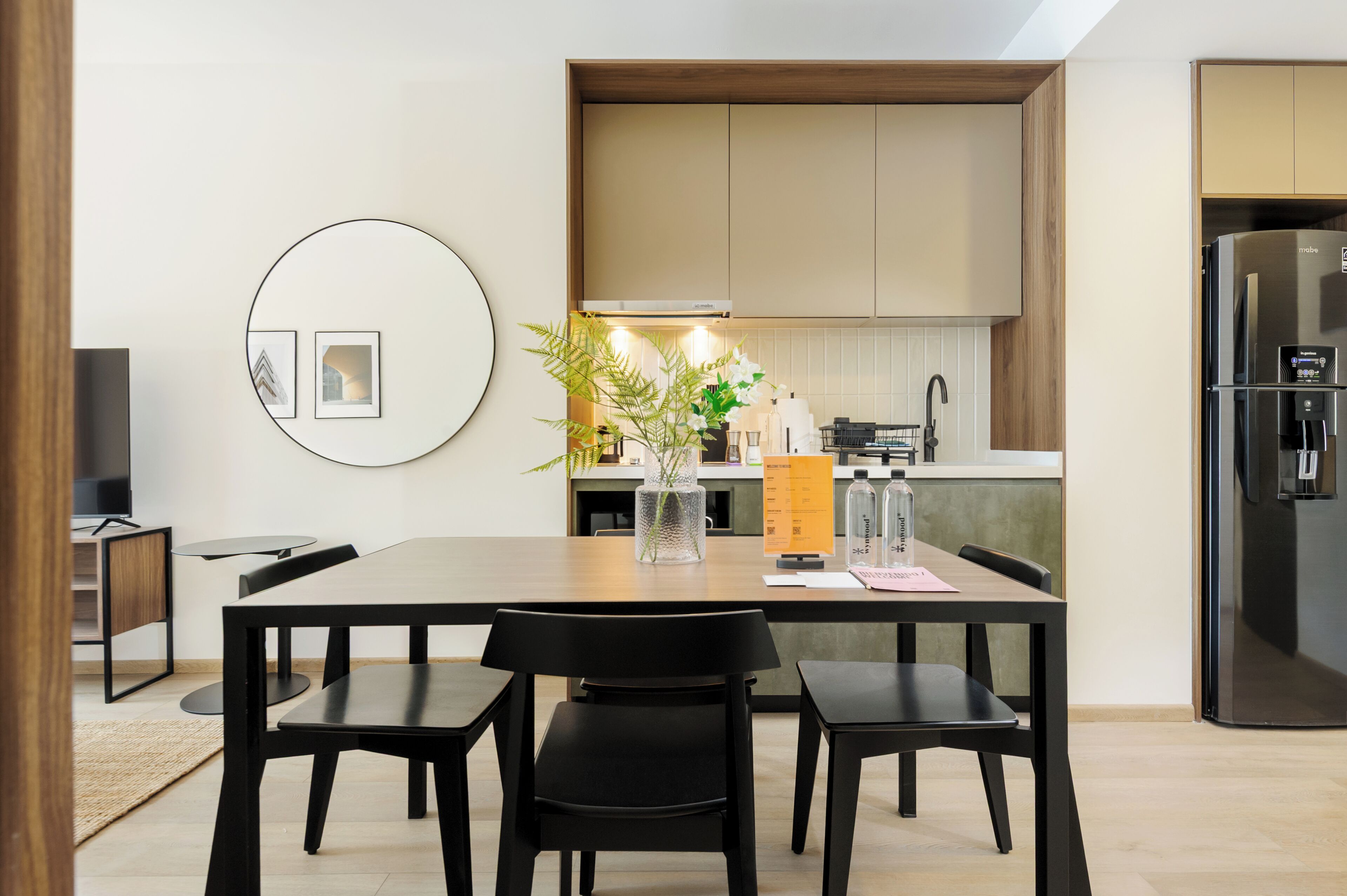 A modern kitchen with wooden cabinets, a built-in oven, and a refrigerator. There is a dining table with chairs and a vase with flowers.