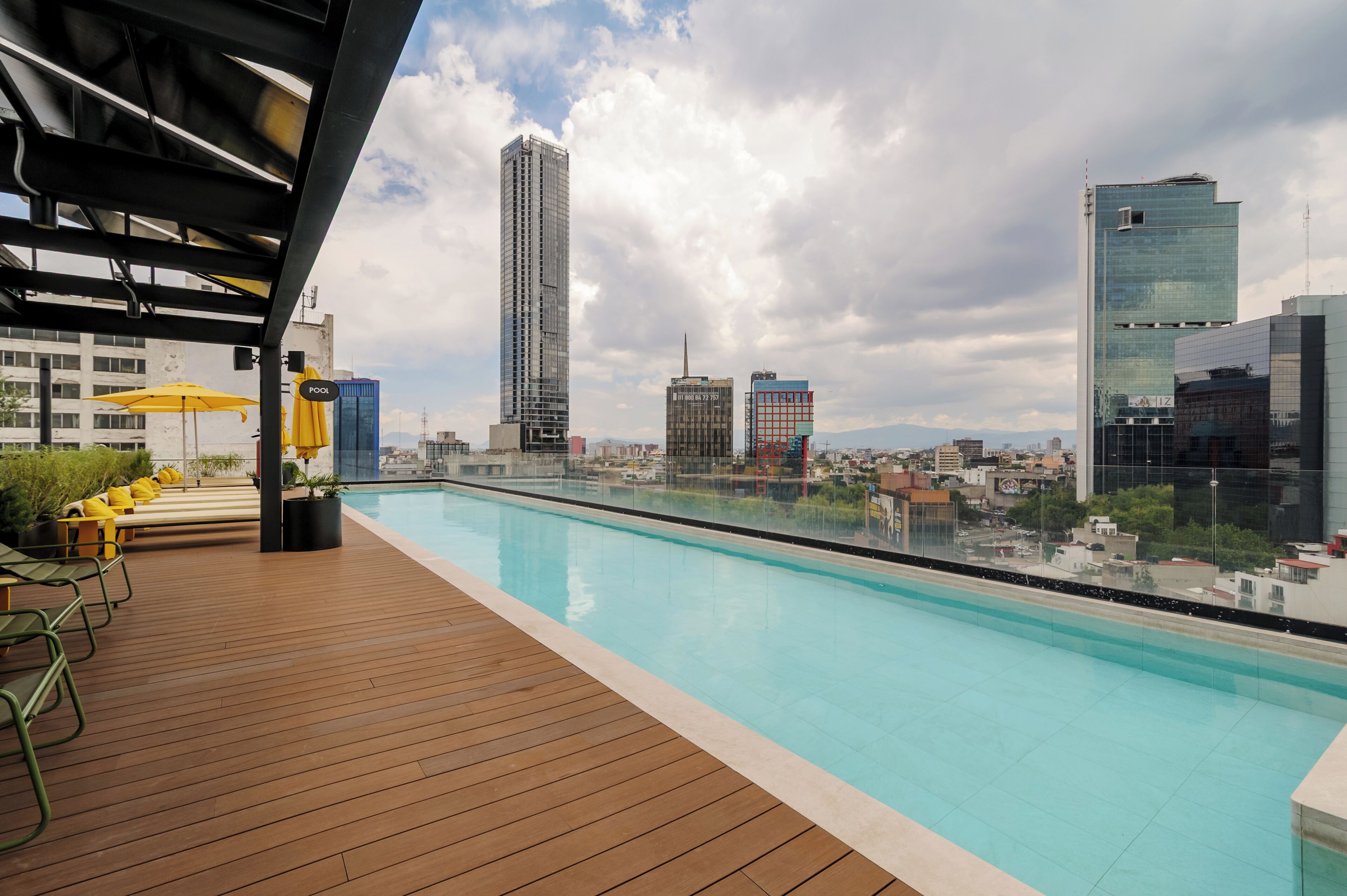 Una piscina en la azotea con vistas al horizonte urbano, terraza de madera y sillones reclinables amarillos.