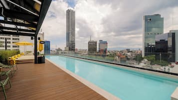 Una piscina en la azotea con vistas al horizonte urbano, terraza de madera y sillones reclinables amarillos.