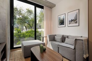 A modern living room with a grey sofa, a wooden coffee table, and a cactus. There are two framed pictures on the wall and a large window with a view of greenery and buildings. - Sullivan by Wynwood House (Mexico City)
