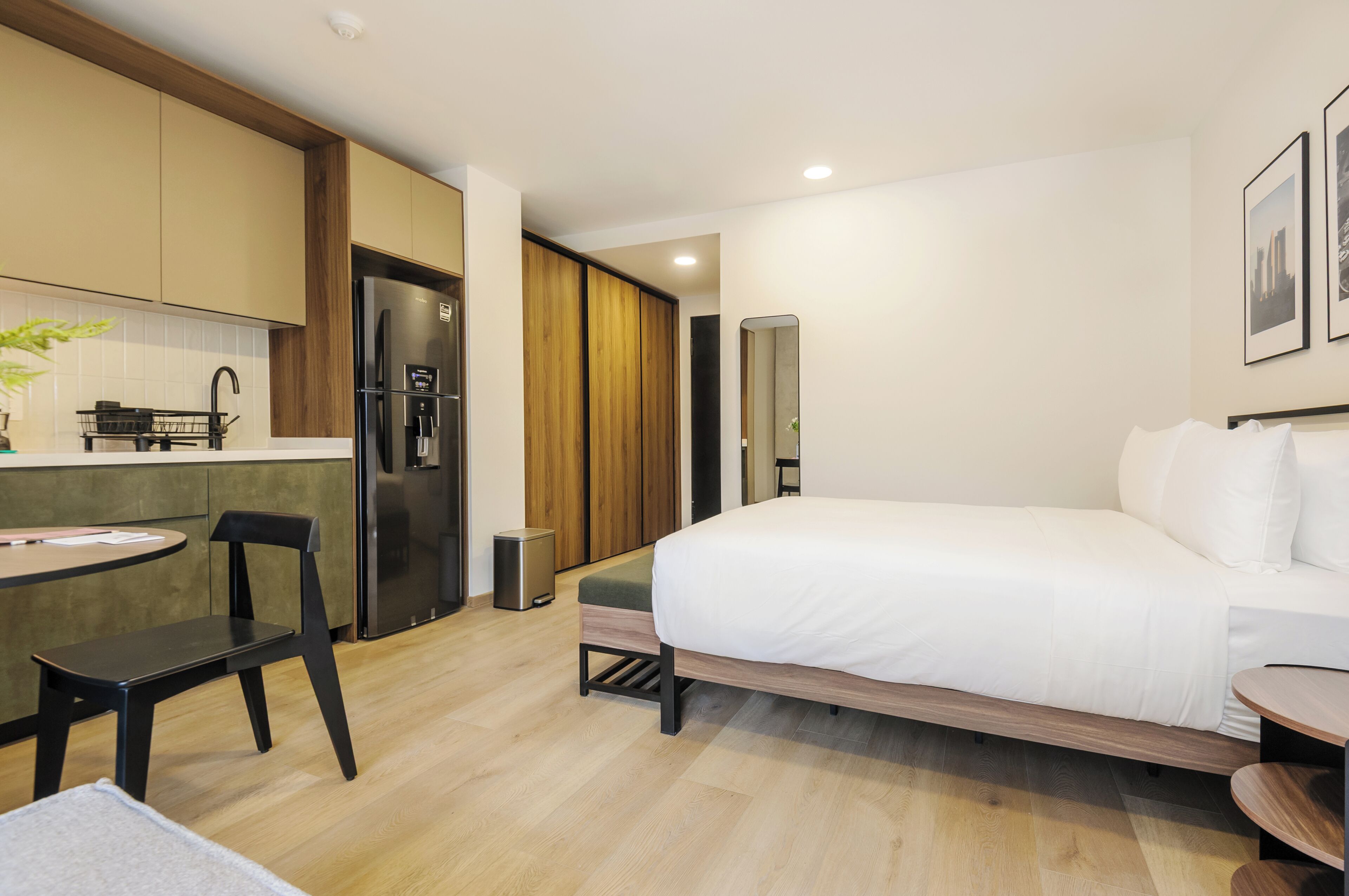 A modern hotel room with a bed, a small desk, a chair, a kitchen area, and a trash bin.