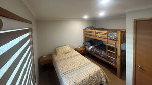 Een kleine kamer met een enkel bed en een houten stapelbed langs de muur, een nachtkastje met een lamp, en een deur aan de rechterkant.