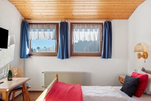 Una camera da letto con soffitto in legno, due finestre con tende, un letto con cuscini, un comodino, un termosifone e una TV fissata al muro.