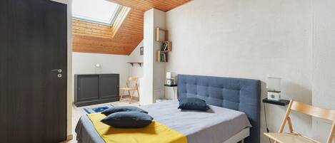 Un dormitorio con cama, mesitas de noche, una silla, un escritorio, un armario, un tragaluz y vistas de la habitación contigua.