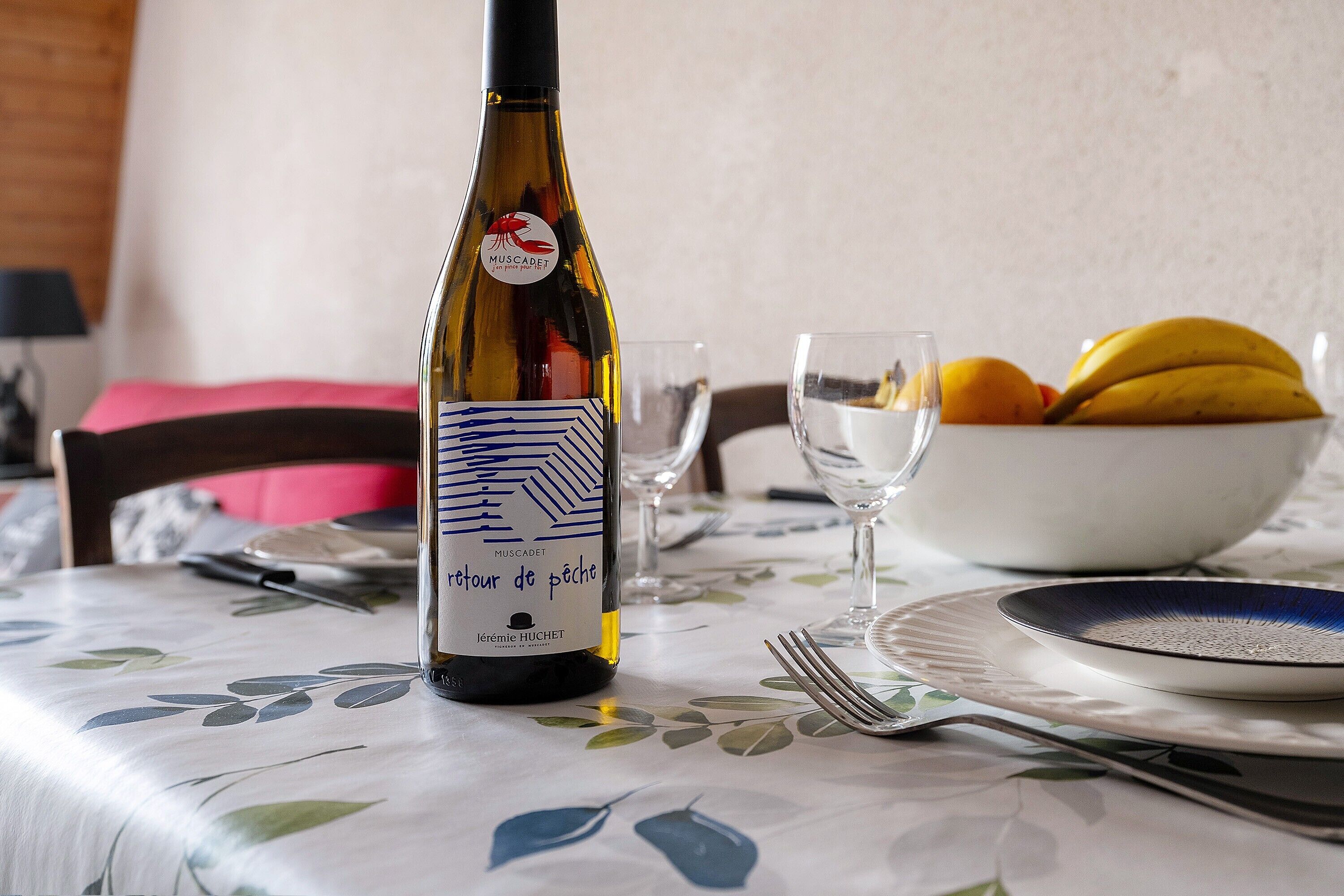 A bottle of Muscadet wine, a bowl of fruit, and a dining table set with plates and cutlery.