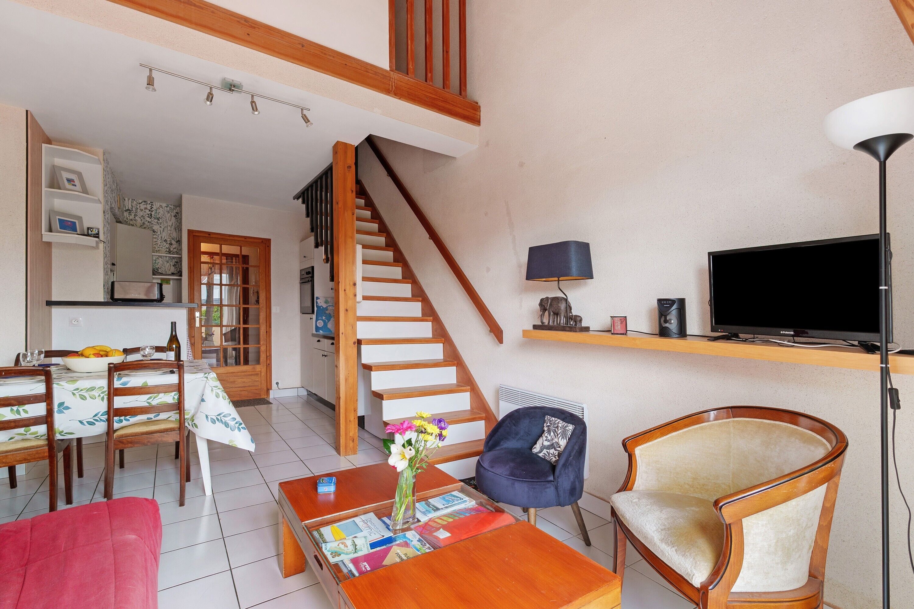 Un salon avec un escalier en bois, un coin repas avec une table et des chaises, un téléviseur posé sur une étagère et un canapé rouge.