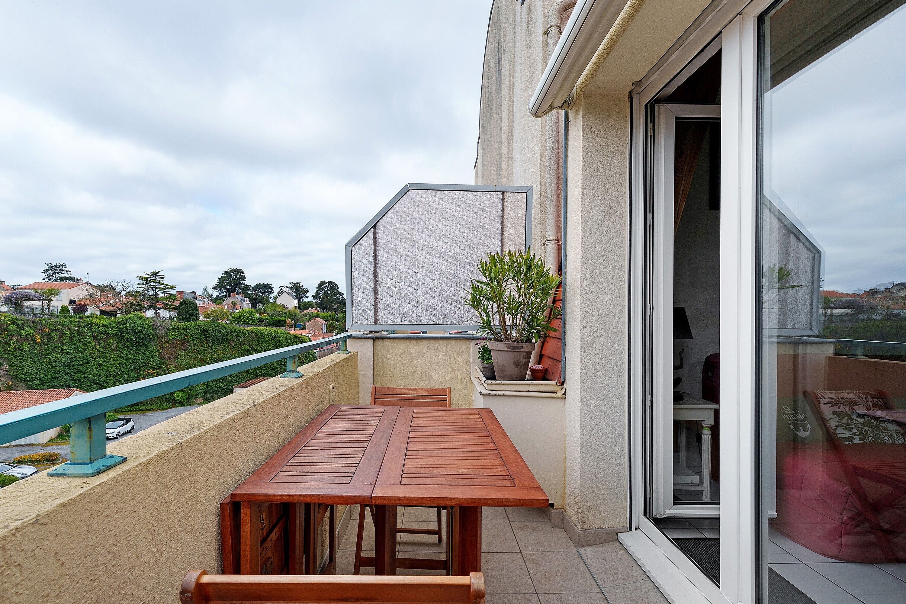 A balcony with a wooden table and chairs, a potted plant, and a view of the neighborhood.