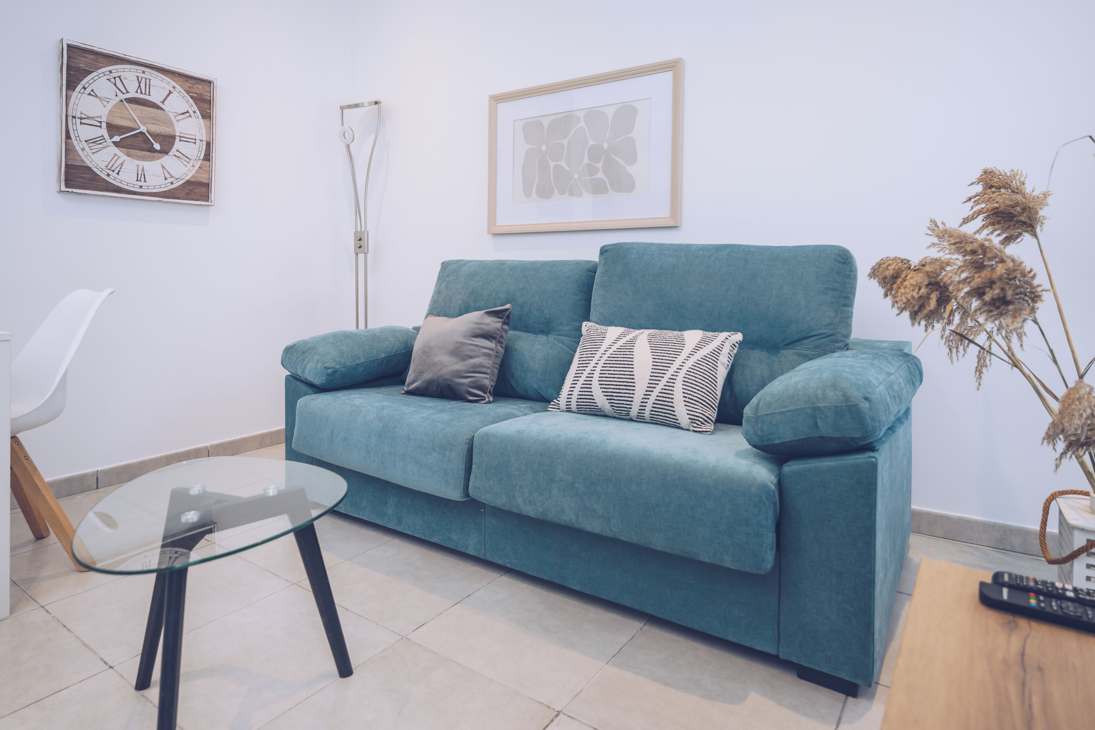 A teal sofa with cushions, a glass table, a framed clock, and a framed abstract art piece on the wall.